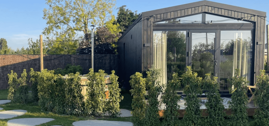 Moderne mantelzorgwoning in een groene tuin, omringd door jonge struiken, met een houten gevel en grote glazen schuifdeuren die uitkijken op een verzorgde buitenruimte.