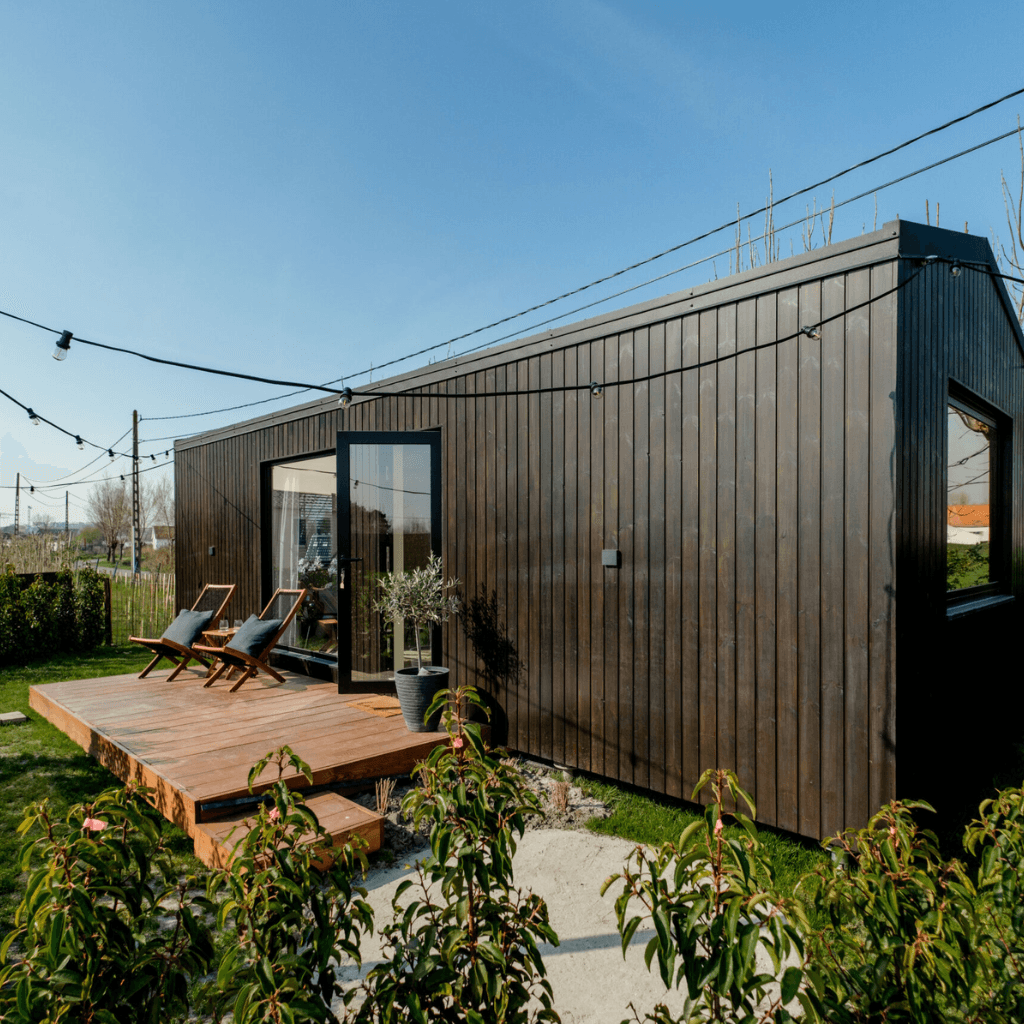 Een vrijstaande jongerenwoning met een moderne houten gevel, omringd door groen, met een uitnodigend terras en zonnige ligstoelen.