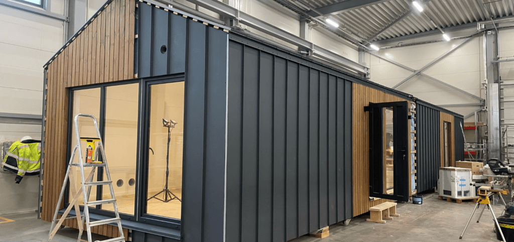 Moderne prefab mantelzorgwoning in aanbouw in een productieruimte, met zwarte gevelpanelen en houten accenten – compacte zorgunit in ontwikkeling